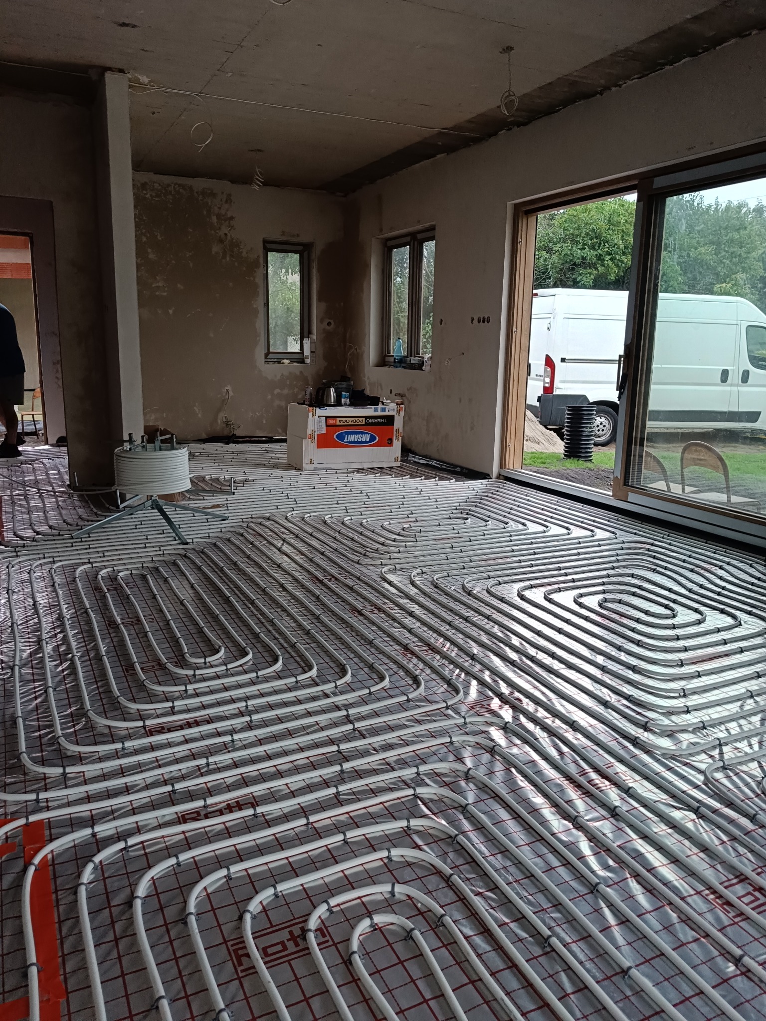 Białe rury ogrzewania podłogowego ułożone spiralnie na folii w surowym wnętrzu. Widoczne okno z widokiem na zewnątrz i biały samochód dostawczy.