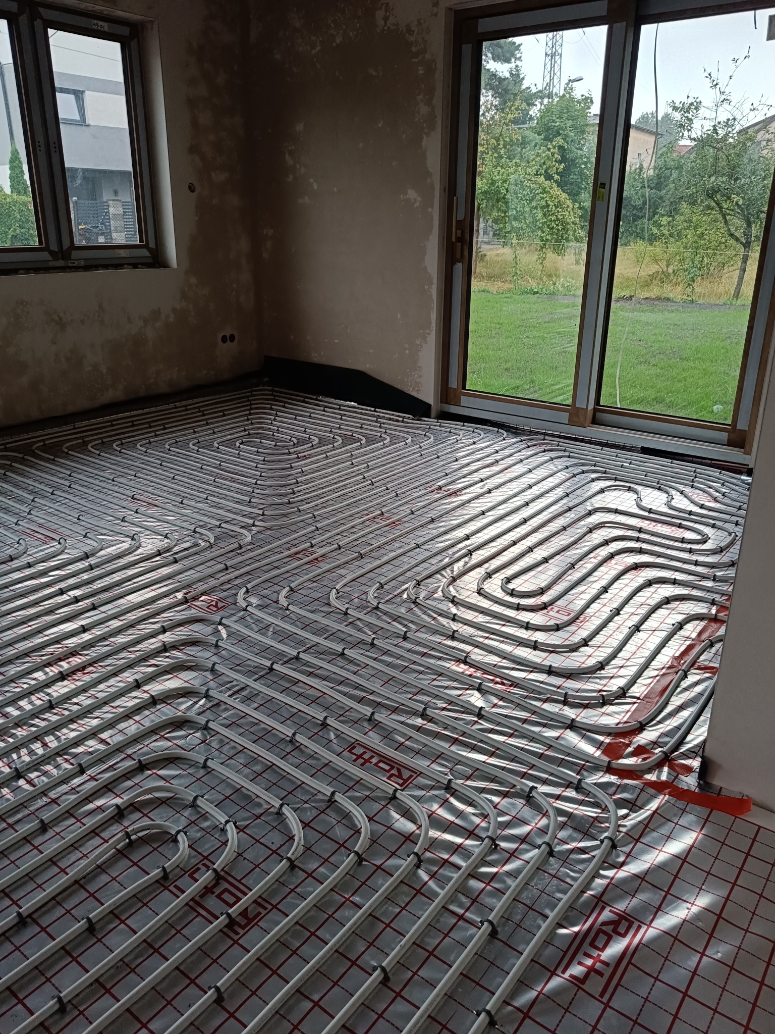 Ułożone rury ogrzewania podłogowego na folii z rastrem w surowym wnętrzu z oknami. Widok z góry, rury ułożone spiralnie, gotowe do zalania wylewką.