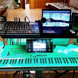 Sebrjan Musik - Stanowisko DJ-a z mikserem, laptopem i keyboardem Yamaha, oświetlone na zielono, na tle sali bankietowej z nakrytymi stołami. Scena gotowa do występu.