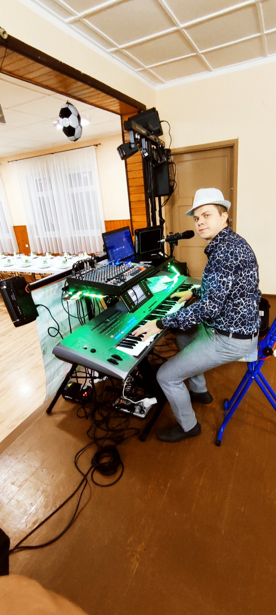 Muzyk w kapeluszu gra na keyboardzie z zielonym podświetleniem, obok mikser i laptop. W tle sala z nakrytymi stołami i oświetlenie sceniczne.