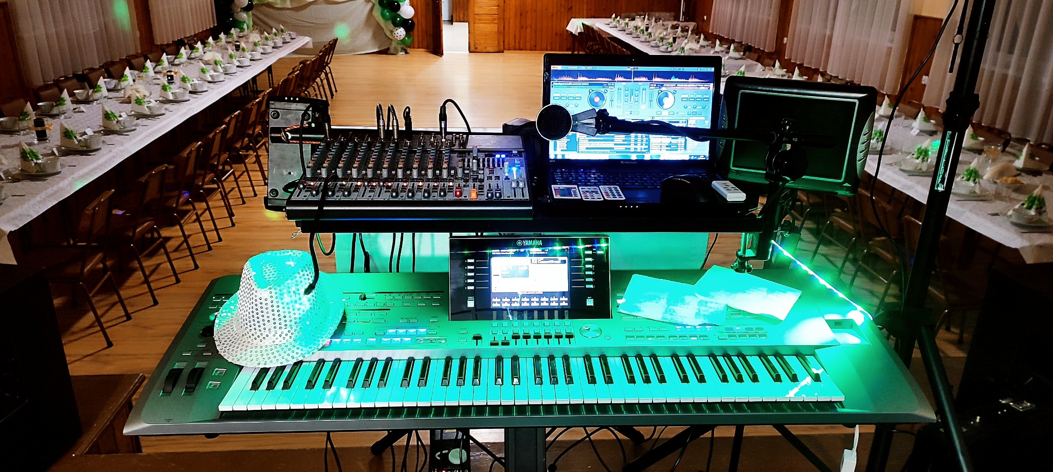 Stanowisko DJ-a z mikserem, laptopem i keyboardem Yamaha, oświetlone na zielono, na tle sali bankietowej z nakrytymi stołami. Scena gotowa do występu.