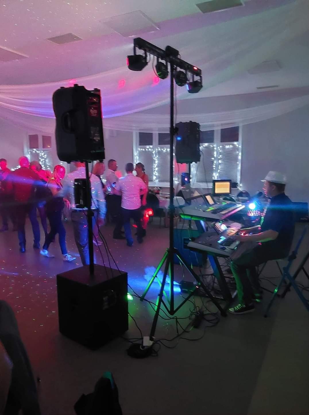 Muzyk w białym kapeluszu gra na keyboardzie podczas imprezy. W tle tańczący ludzie, oświetlenie sceniczne i dekoracje.