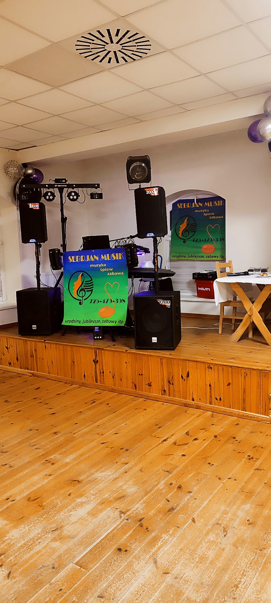Scena z nagłośnieniem i oświetleniem, baner reklamowy zespołu muzycznego 'Sebrjan Musik' na drewnianym podeście. Widoczne głośniki, reflektory i stolik.