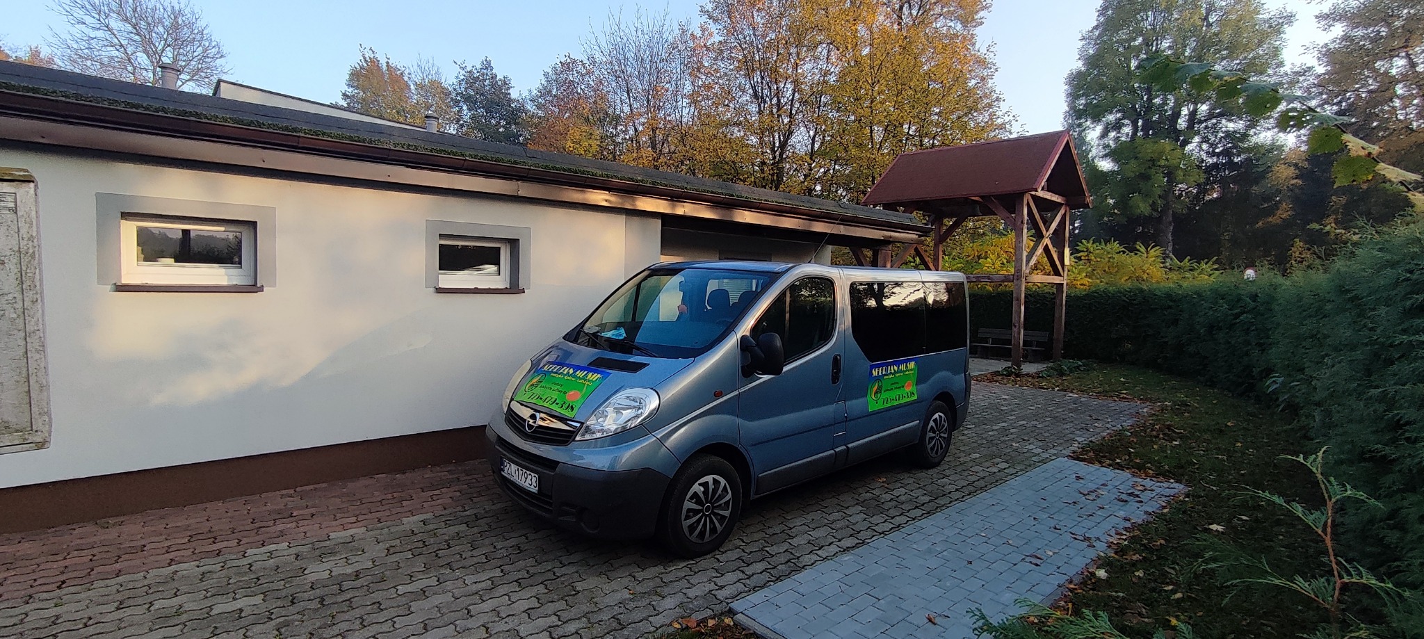 Błękitny Opel Vivaro z naklejką 'Serbian Musik' parkuje na brukowanym podjeździe przed budynkiem i drewnianą altaną. Jesienne drzewa w tle.