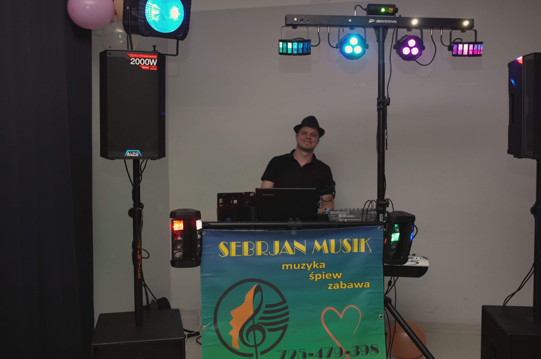 DJ z uśmiechem za konsoletą, nagłośnienie i oświetlenie sceniczne. Baner z napisem 'Sebrjan Musik muzyka śpiew zabawa'.