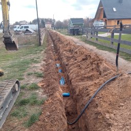 Mix-Kop - Długi wykop w ziemi z widocznymi niebieskimi rurami i kablem, obok stoi koparka. W tle domy i ogrodzenie. Prace ziemne w toku.