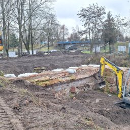 Mix-Kop - Żółta koparka JCB przy wykopie z ceglanym murem i workami z piaskiem. W tle ziemia, drzewa i most. Prace ziemne w pochmurny dzień.