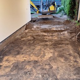 Mix-Kop - Żółta mini koparka na wyrównanym terenie pod zadaszeniem, w tle wywrotka z ziemią i zielony żywopłot. Przygotowanie terenu pod budowę.