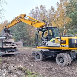 Mix-Kop - Żółta koparka JCB JS160W ładuje gruz do skrzyni ciężarówki na placu budowy, widoczne ślady opon na ziemi i jesienne drzewa w tle. Prace ziemne w toku.