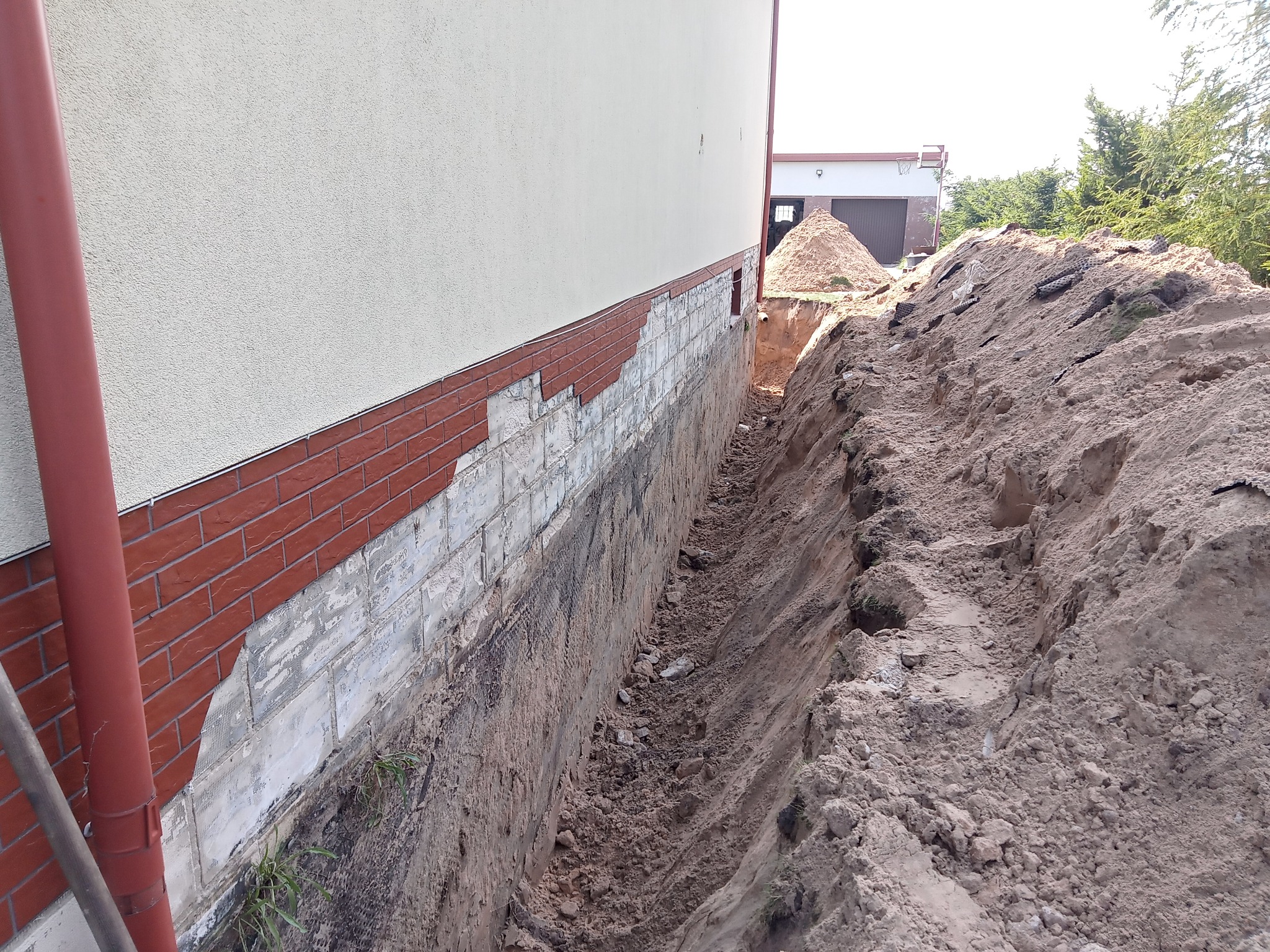 Wykop fundamentów w Pisz, widoczny rów wzdłuż ściany budynku z odsłoniętą cegłą i stertą ziemi w tle. Prace ziemne przygotowawcze pod fundament.