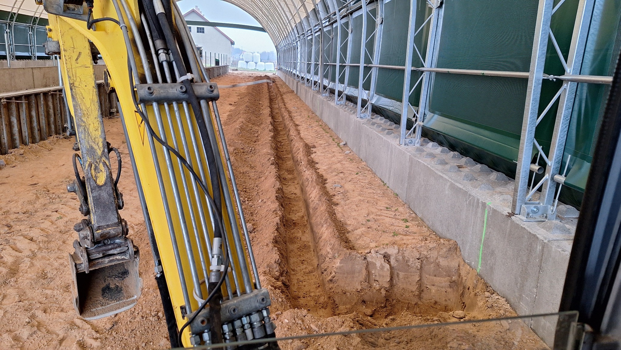 Żółta koparka w trakcie wykonywania wykopu pod fundamenty wewnątrz hali. Widoczne równe rowy w ziemi i konstrukcja hali w tle. Prace ziemne w toku.