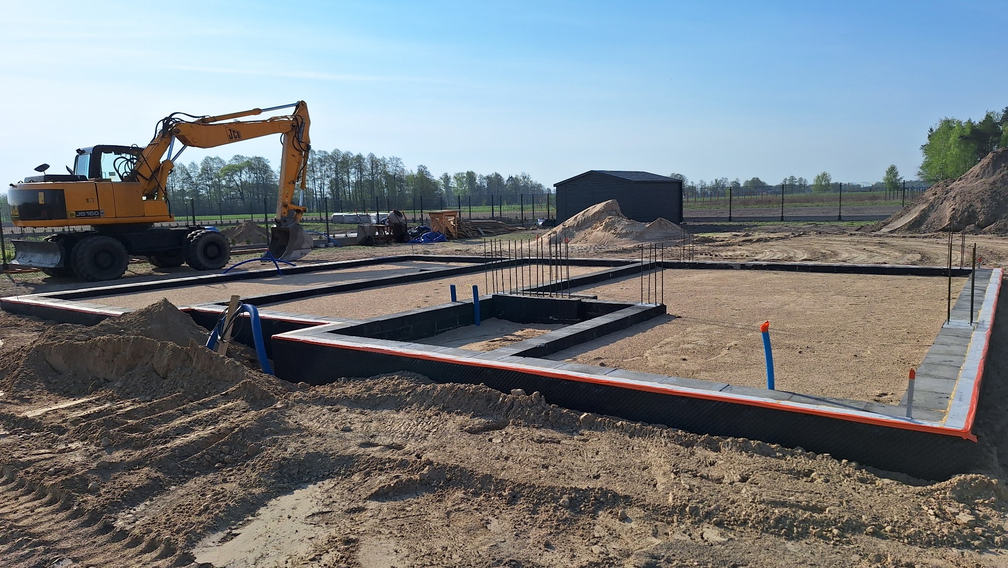 Żółta koparka JCB obok przygotowanego zbrojenia fundamentów z izolacją, na piaszczystym terenie budowy w słoneczny dzień. Widoczny barak i hałda ziemi.