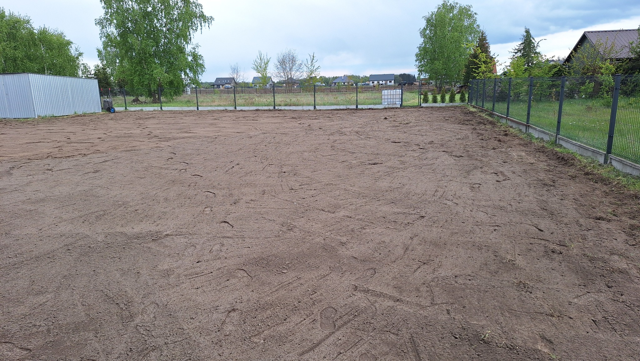Przygotowany teren pod trawnik z rolki, widoczne ślady po maszynach, ogrodzenie panelowe, budynki w tle. Teren wyrównany, gotowy do położenia trawy.