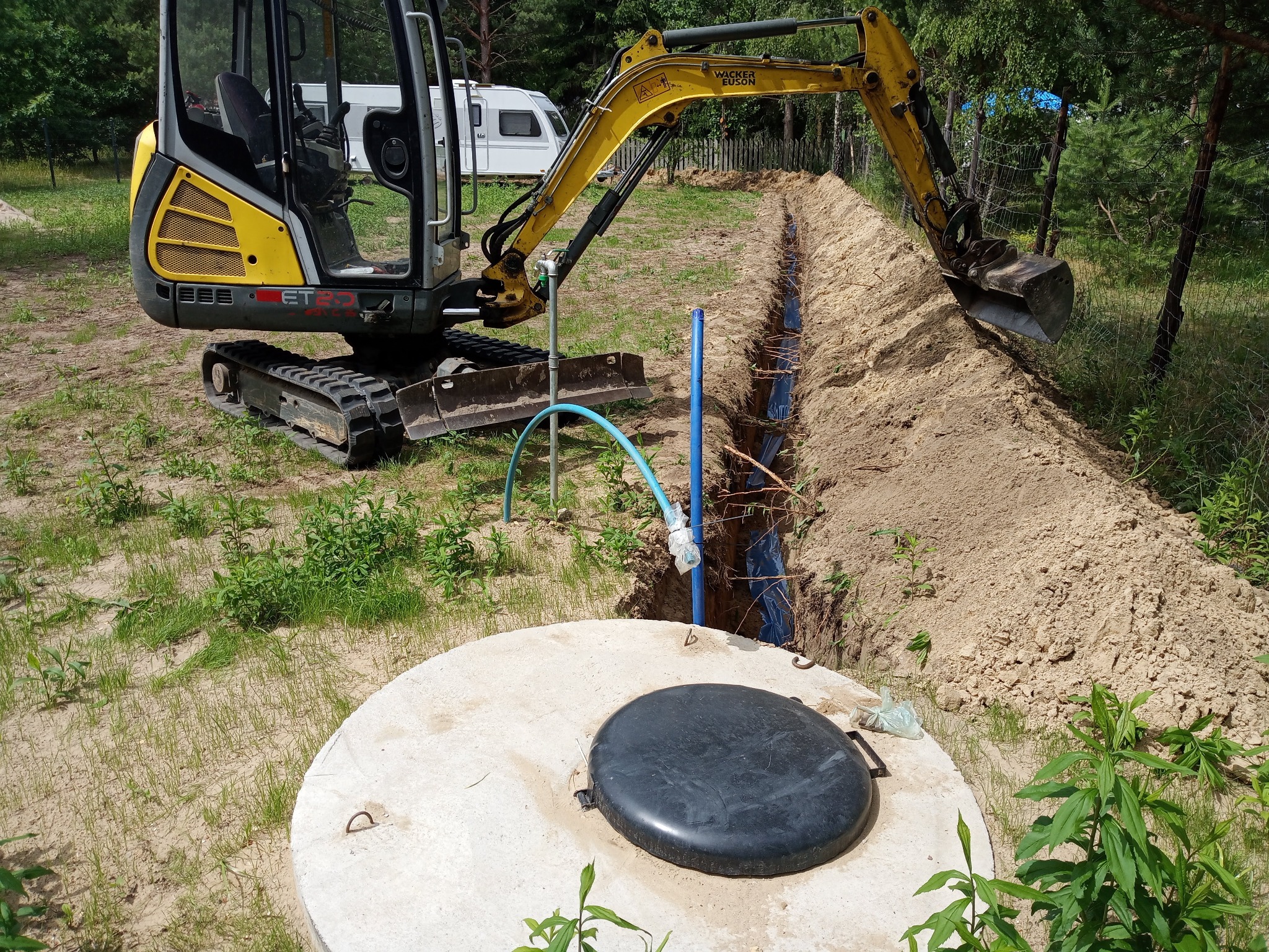Żółty minikoparka Wacker Neuson obok betonowego szamba z czarną pokrywą, w tle wykopany rów z niebieskimi rurami. Widoczne drzewa i przyczepa kempingowa.