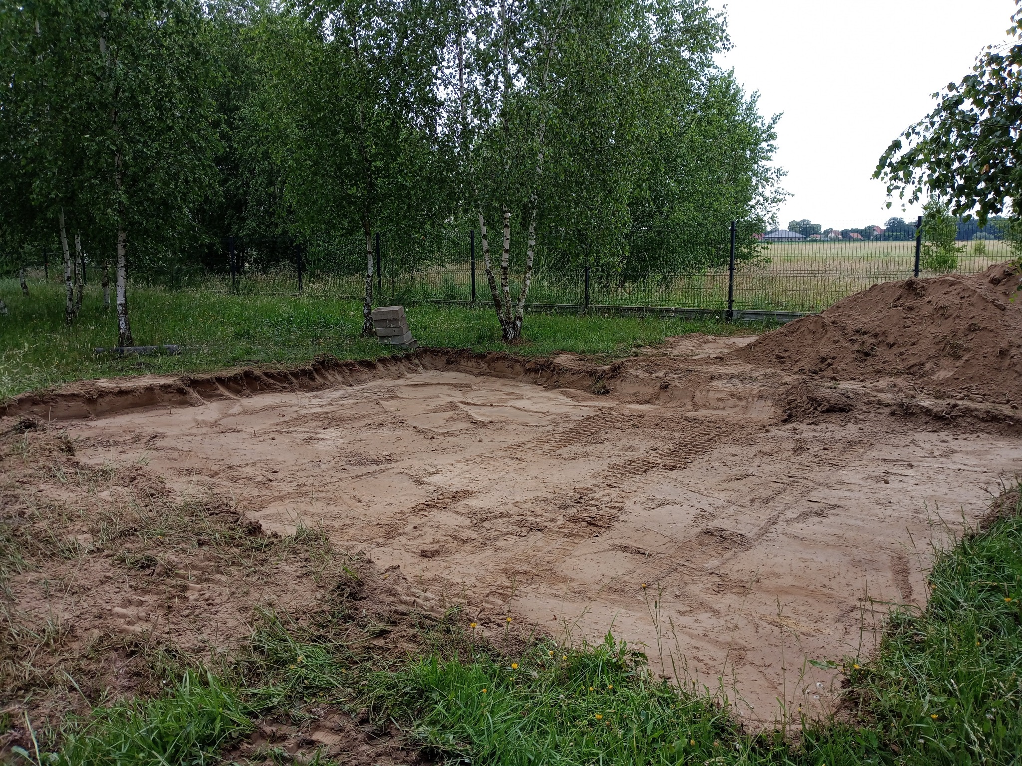 Wyrównany teren pod budowę z widocznymi śladami opon, obok pryzma ziemi i trawa. W tle drzewa i ogrodzenie z siatki.