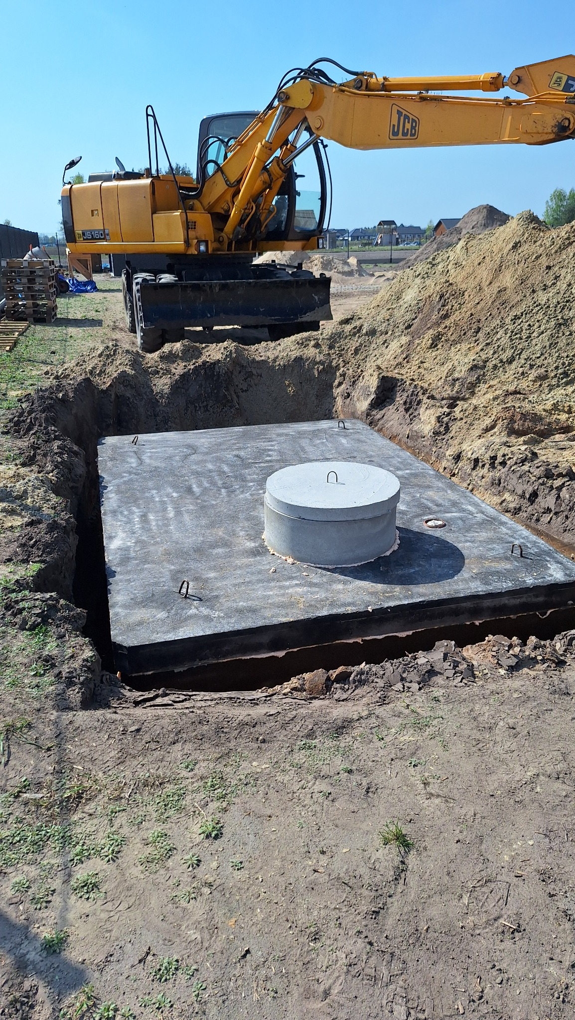 Betonowy zbiornik z pokrywą w wykopie, wspierany przez koparkę JCB. Widoczne ślady prac ziemnych i przygotowany teren pod instalację.