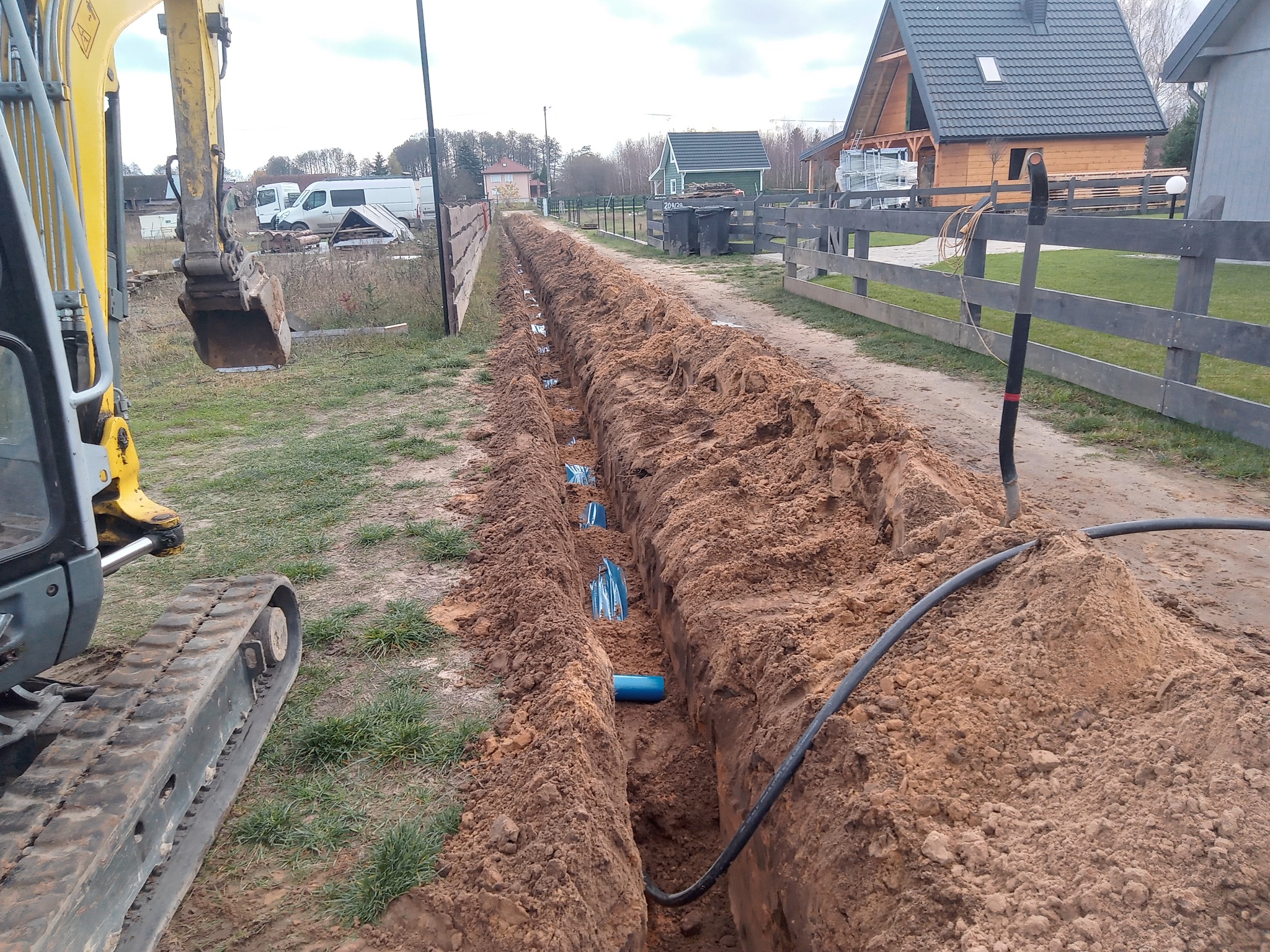Długi wykop w ziemi z widocznymi niebieskimi rurami i kablem, obok stoi koparka. W tle domy i ogrodzenie. Prace ziemne w toku.