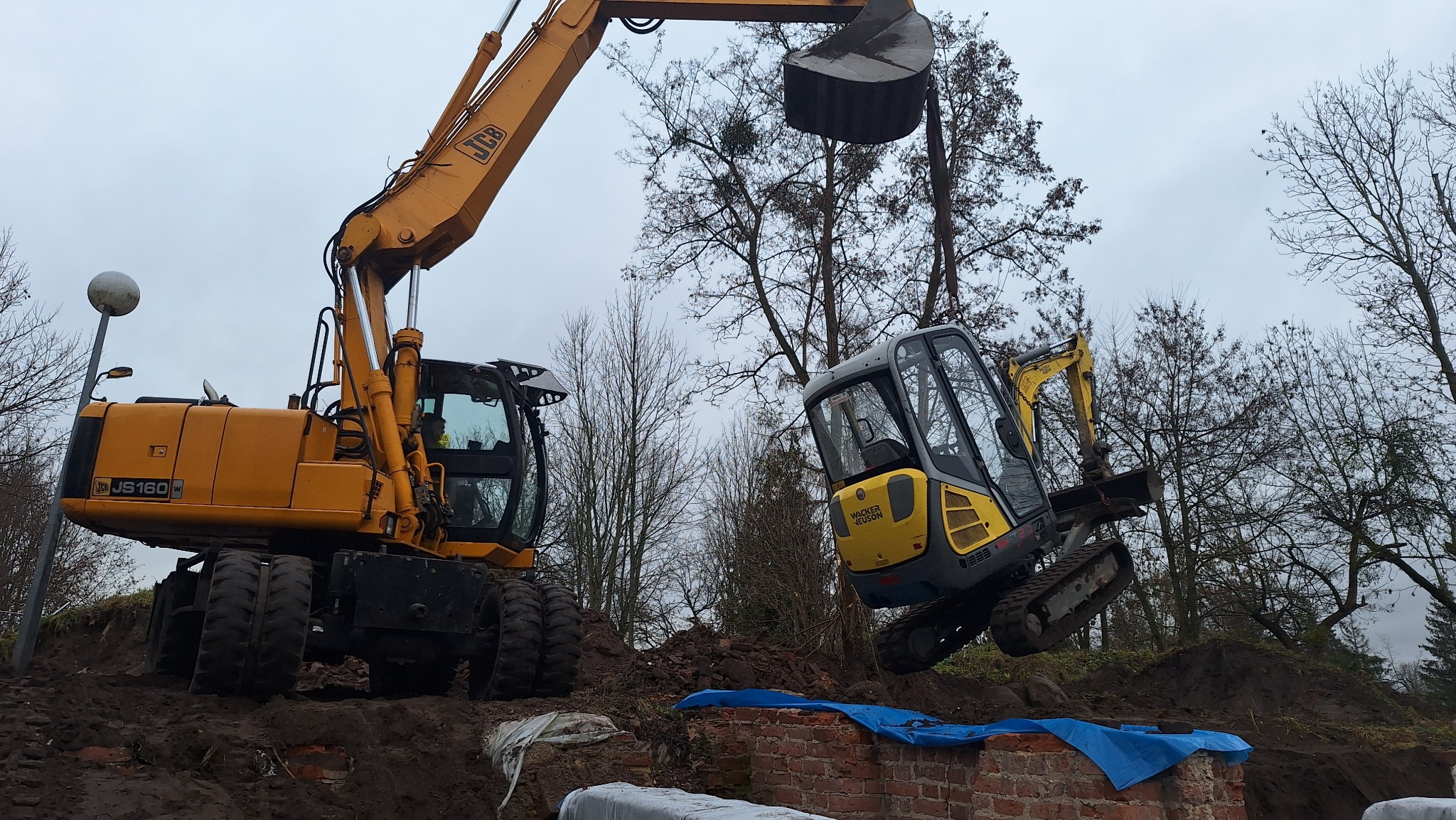Żółta koparka JCB podnosi mniejszą koparkę Wacker Neuson nad wykopem z ceglanymi elementami, na tle drzew i pochmurnego nieba. Prace ziemne w toku.