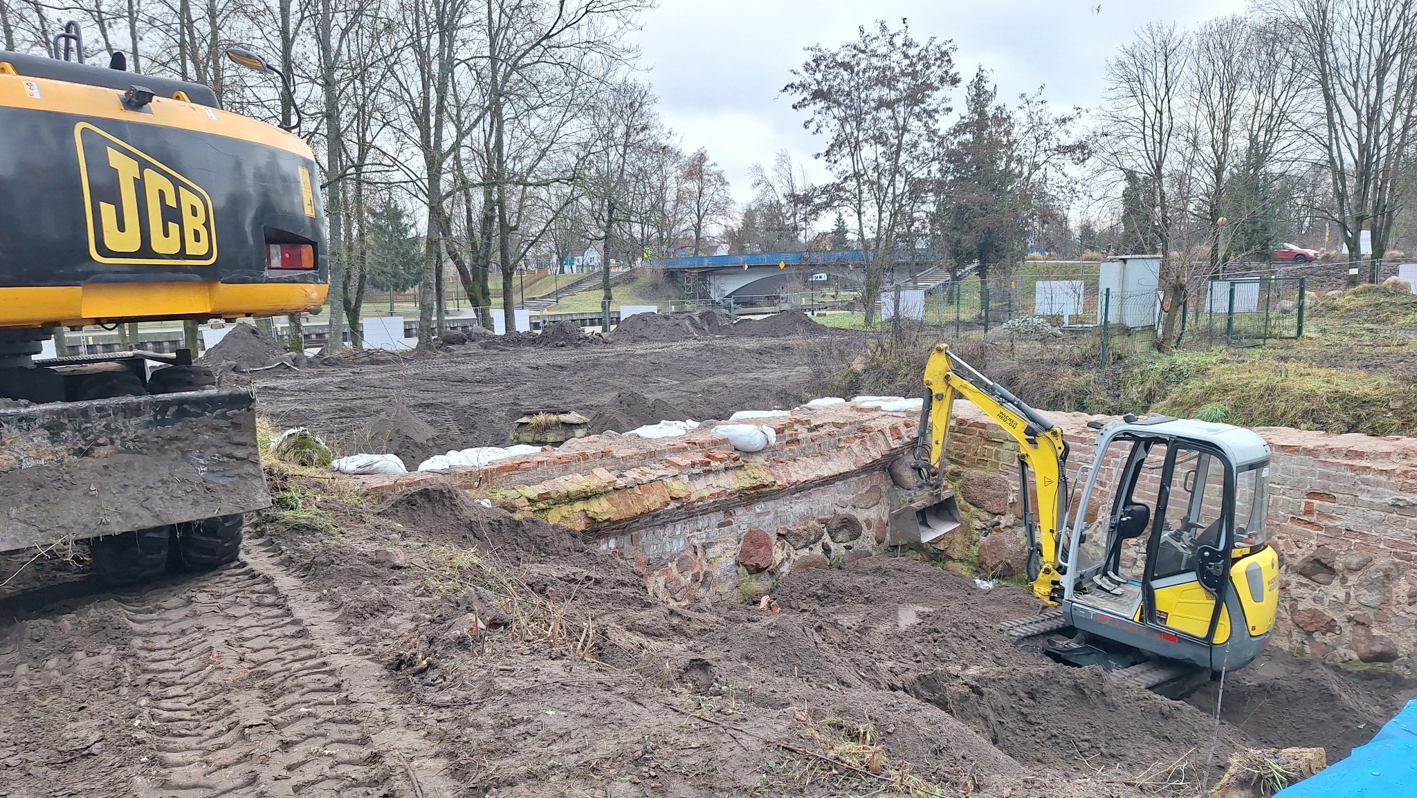 Żółta koparka JCB przy wykopie z ceglanym murem i workami z piaskiem. W tle ziemia, drzewa i most. Prace ziemne w pochmurny dzień.