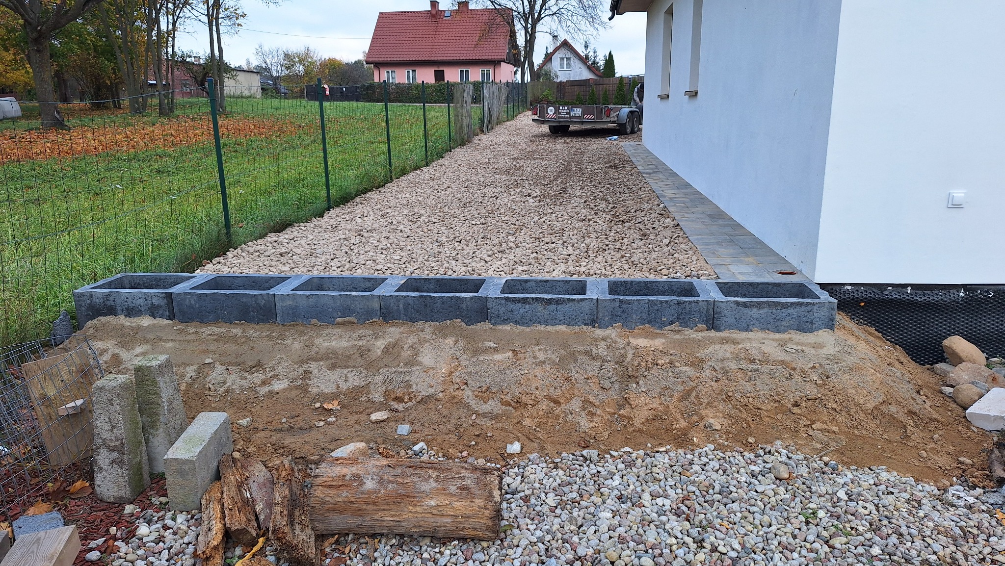 Budowa drenażu: rząd betonowych bloczków, podsypka żwirowa, kostka brukowa przy ścianie budynku. Widoczna trawa i ogrodzenie w tle. Prace ziemne w toku.