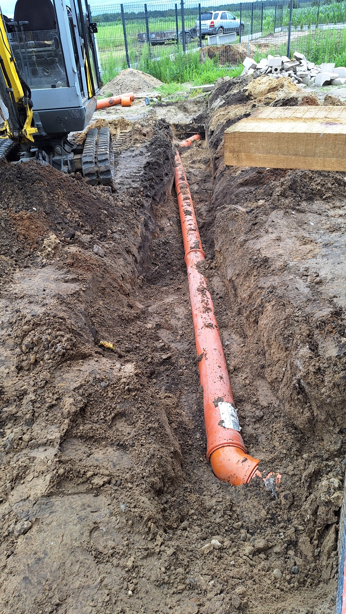Pomarańczowa rura kanalizacyjna ułożona w wykopie ziemnym, widoczna z góry. W tle koparka i ogrodzenie posesji. Etap prac przyłącza wodno-kanalizacyjnego.