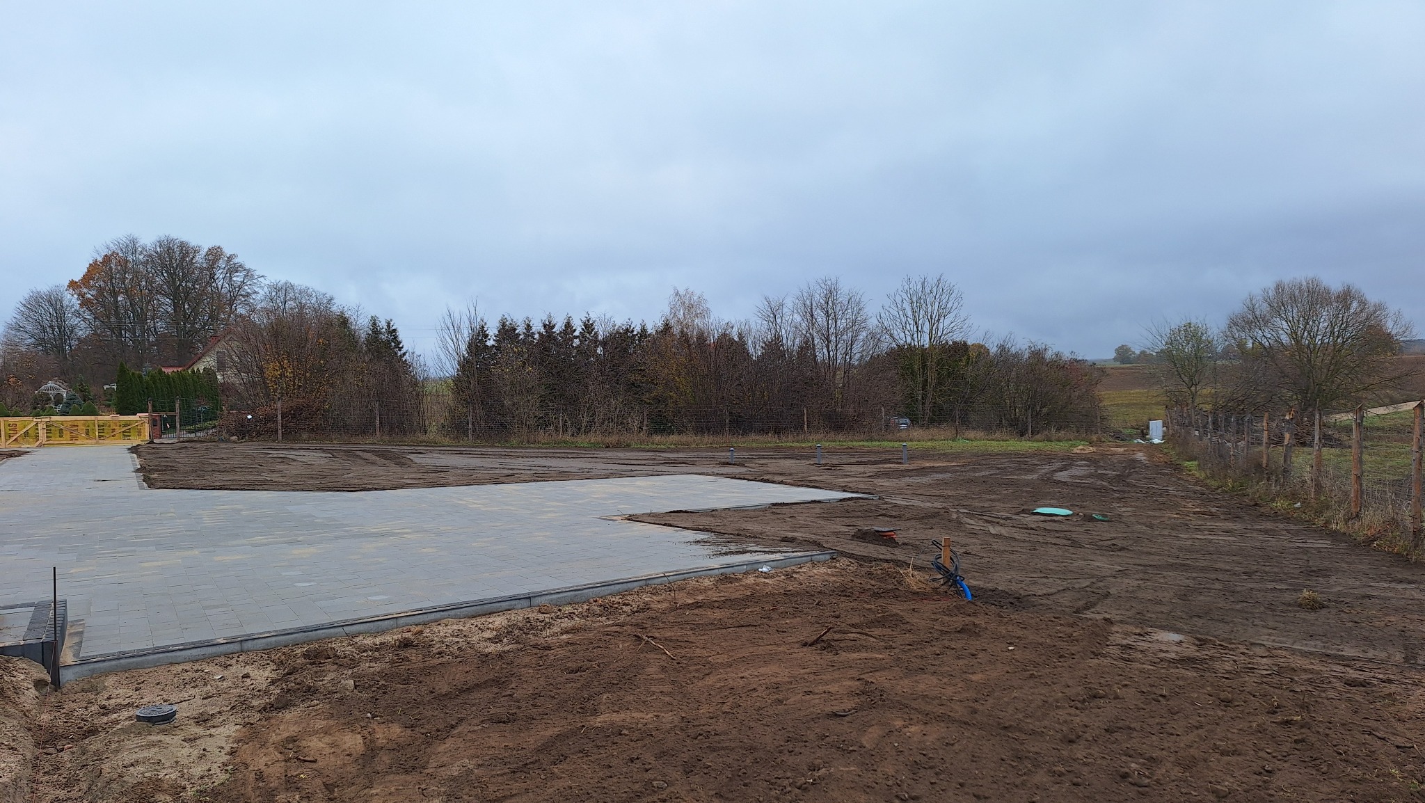 Częściowo ułożona, szara kostka brukowa na przygotowanym terenie, widoczne obrzeża. W tle drzewa i pochmurne niebo. Prace brukarskie w toku, teren wyrównany.