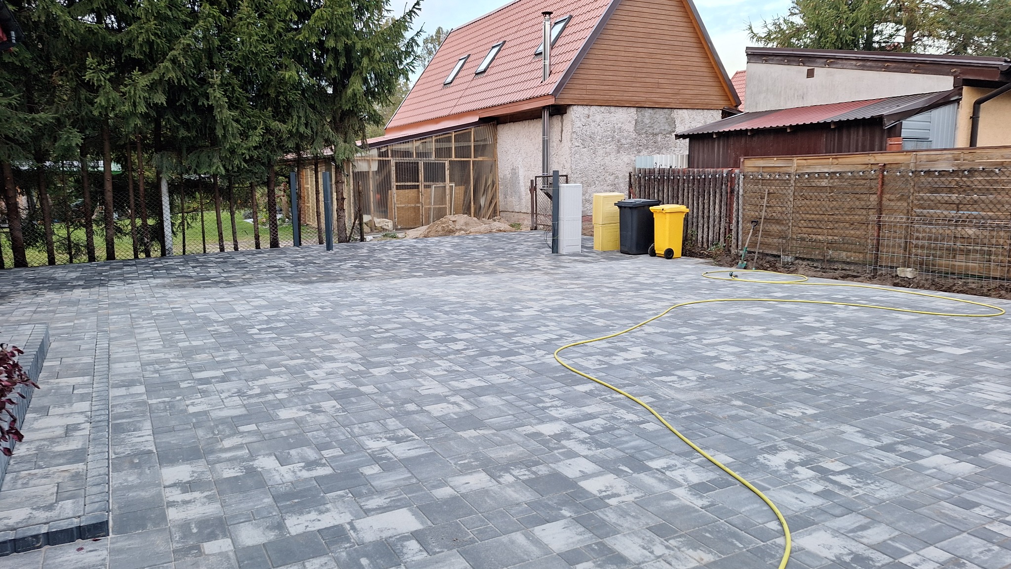 Ułożona kostka brukowa w szaro-białej tonacji na posesji, widoczne pojemniki na śmieci i częściowo ogrodzenie. Żółty wąż ogrodowy leży na kostce.