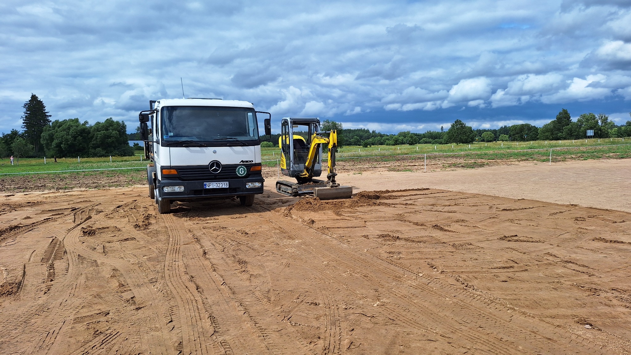 Żółta mini koparka i biała ciężarówka Mercedes na wyrównanym, piaszczystym terenie. W tle zielone drzewa i pochmurne niebo.