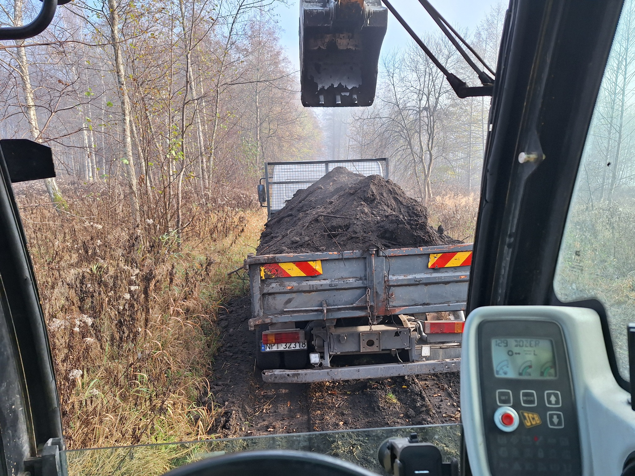 Wywóz ziemi: Widok z kabiny koparki na wywrotkę załadowaną ziemią, w tle drzewa i zarośla. Praca w toku, widoczne błoto na drodze.