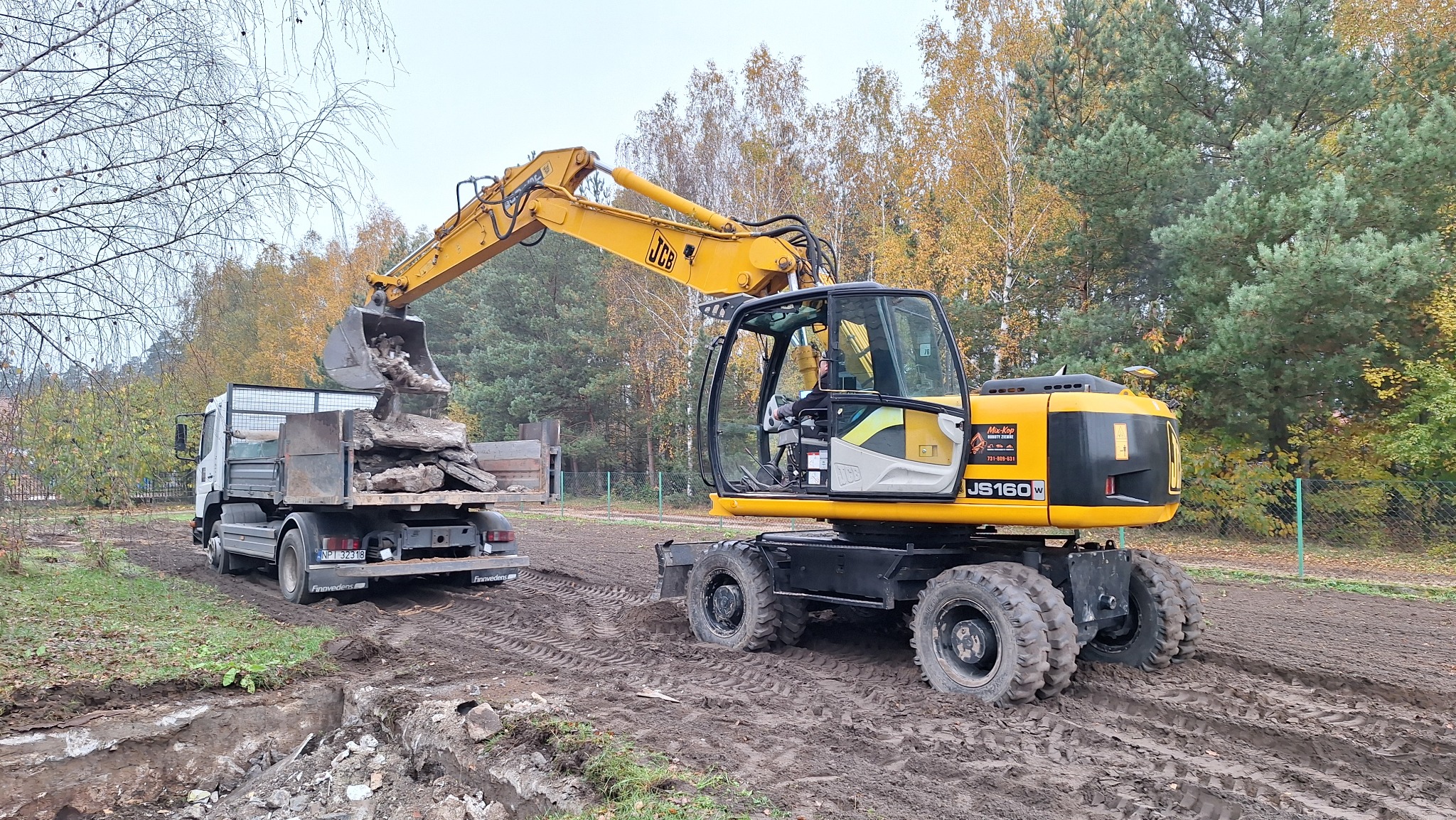 Żółta koparka JCB JS160W ładuje gruz do skrzyni ciężarówki na placu budowy, widoczne ślady opon na ziemi i jesienne drzewa w tle. Prace ziemne w toku.