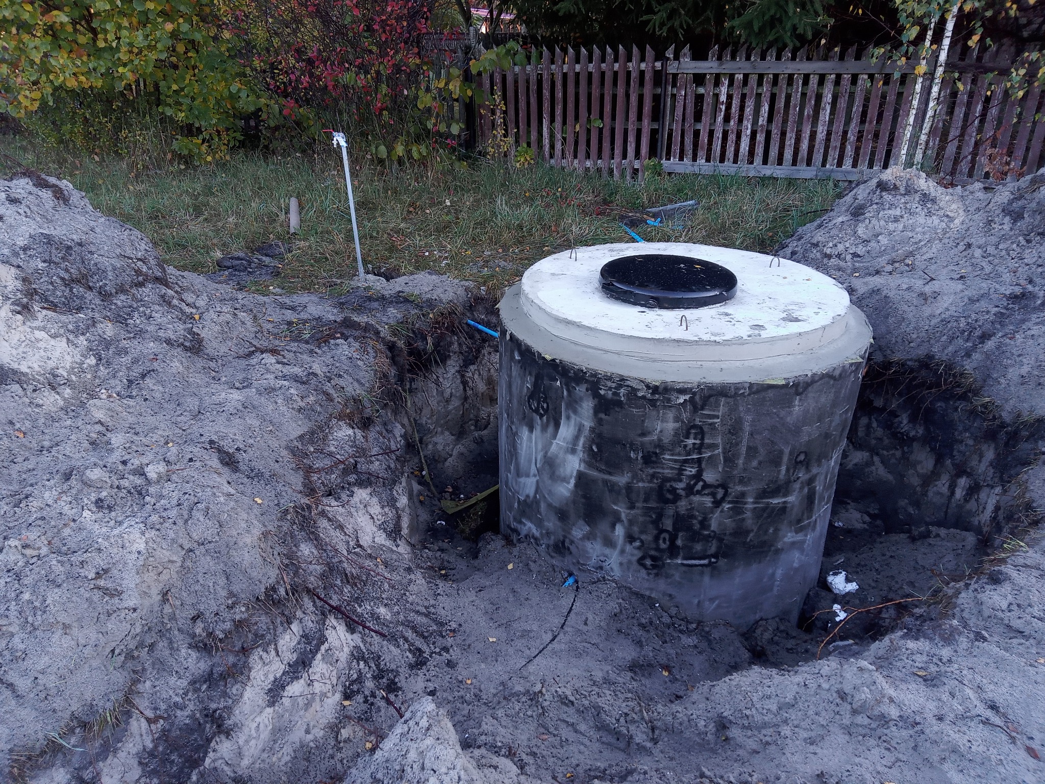 Betonowy zbiornik z czarną pokrywą, częściowo zagłębiony w ziemi, otoczony wykopem. Widoczne przyłącza i elementy instalacji. W tle trawa i drewniane ogrodzenie.