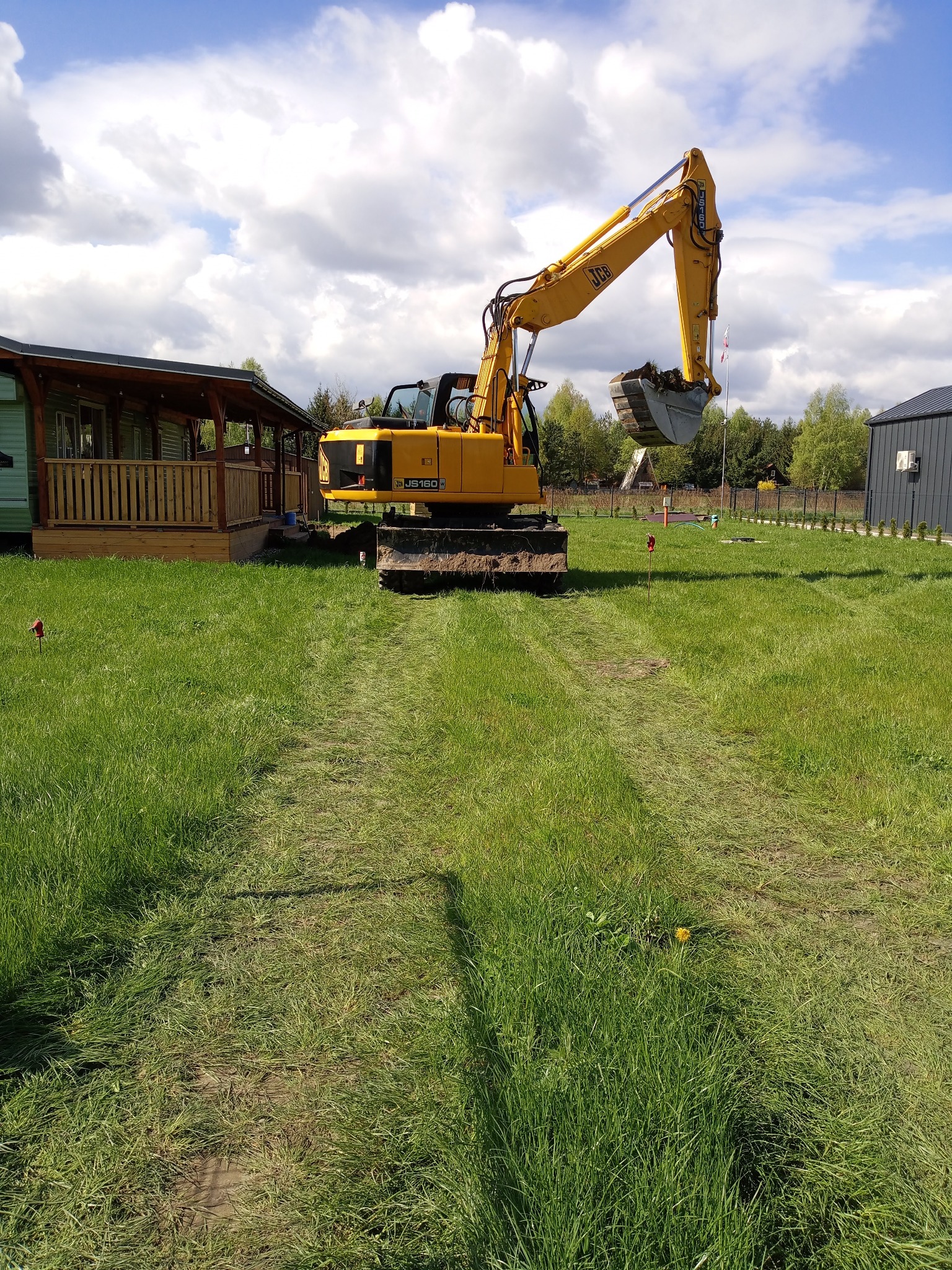 Żółta koparka JCB JS160LC na trawniku, ślady opon w trawie, domki w tle, błękitne niebo z chmurami. Prace ziemne w toku.