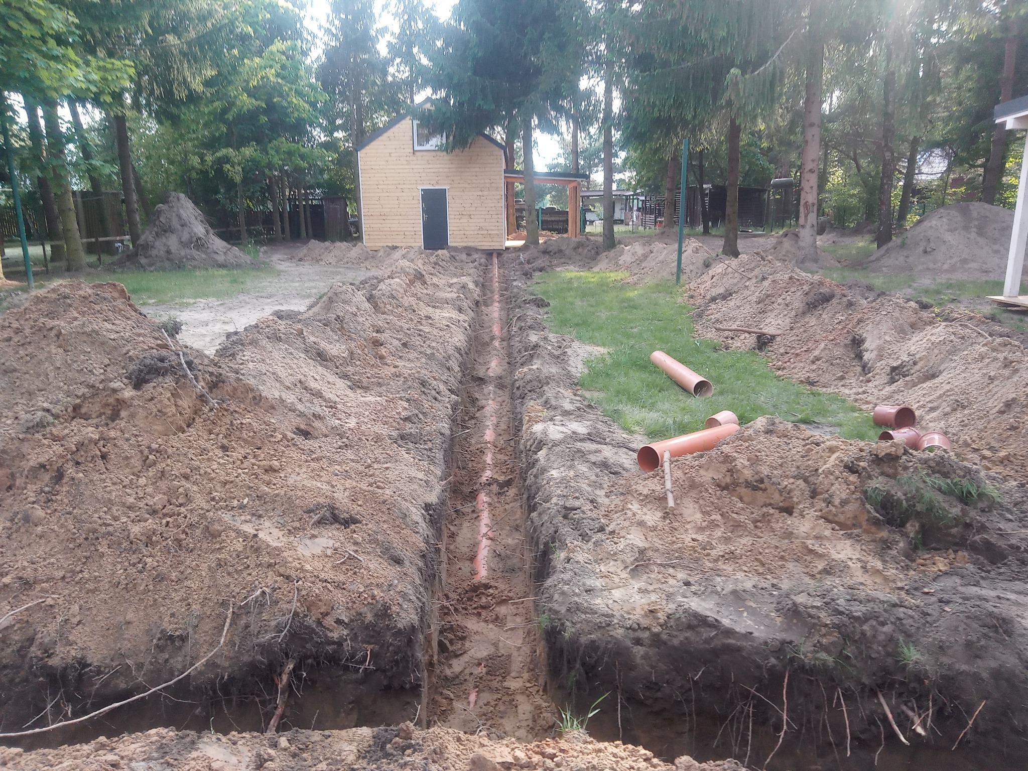 Wykopany rów z ułożonymi rurami kanalizacyjnymi na dnie, widok z góry, w tle mały budynek gospodarczy i drzewa. Prace ziemne w toku.