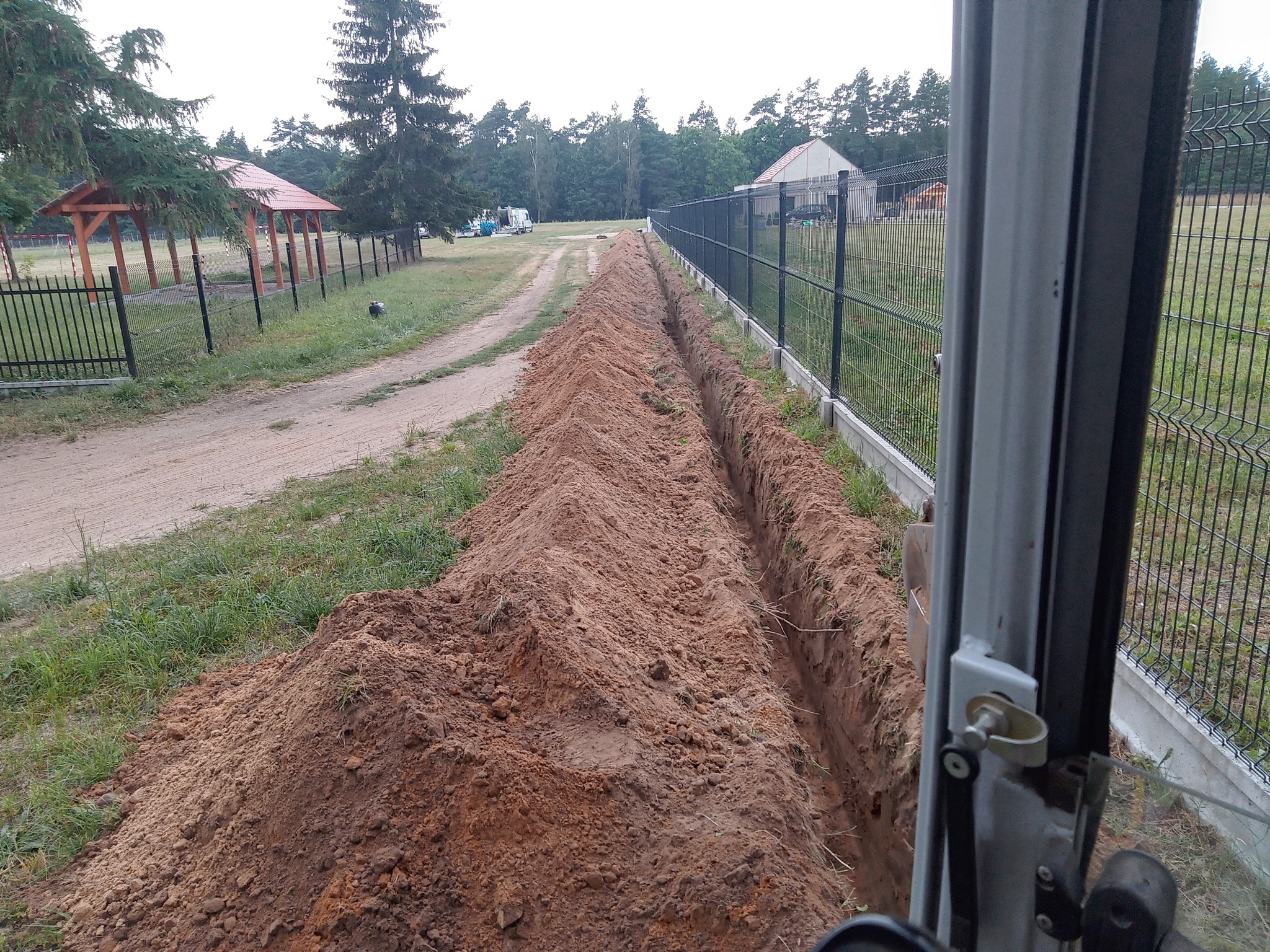 Długi wykop w ziemi, widok z kabiny maszyny budowlanej, obok ogrodzenie z siatki. Świeżo wykopana ziemia usypana wzdłuż wykopu. Prace ziemne w toku.