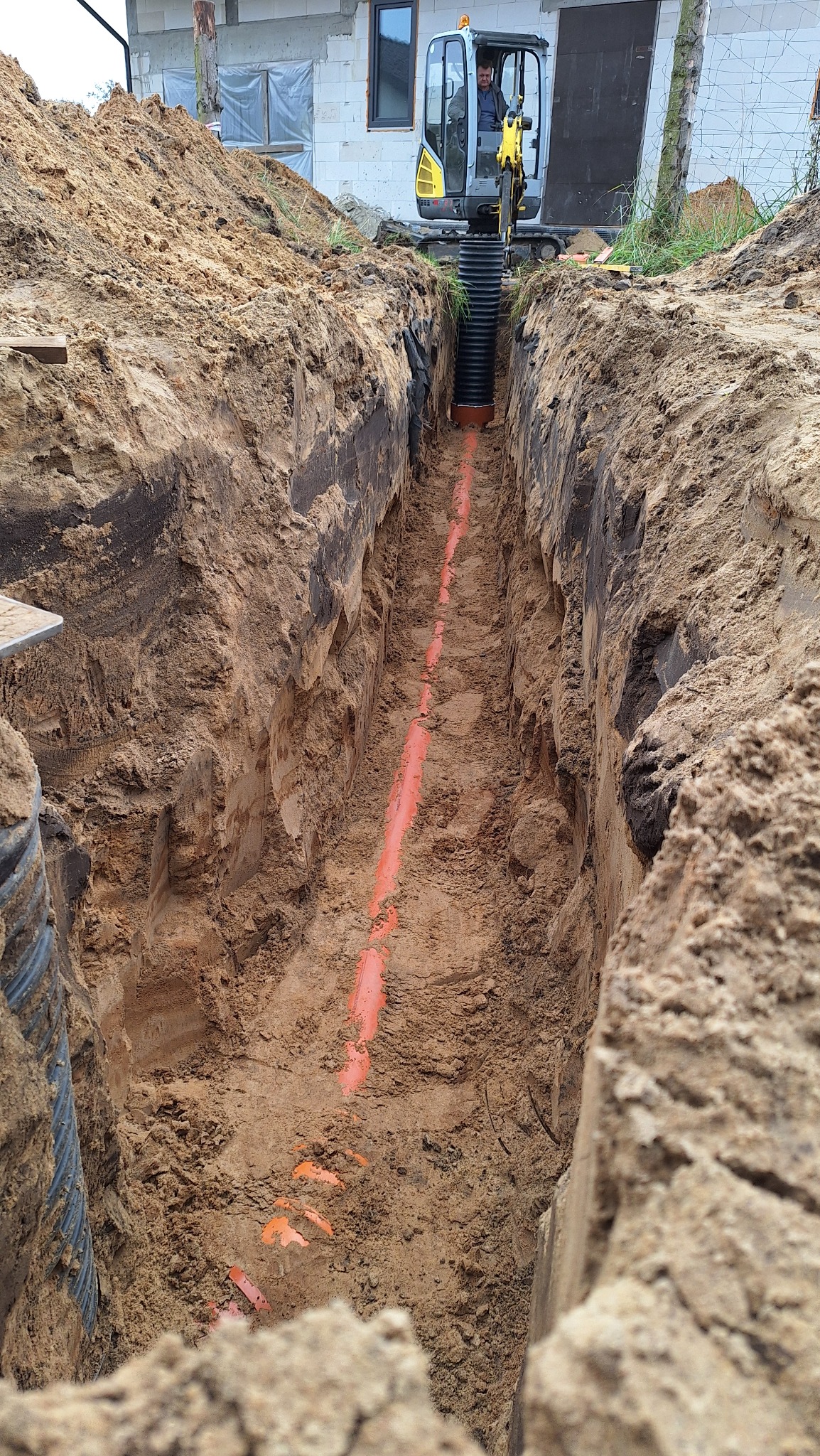 Wykop pod przyłącze wod-kan z pomarańczową rurą na dnie, widoczny operator koparki i fragment budynku w tle. Wykopane ściany z wyraźnymi warstwami ziemi.