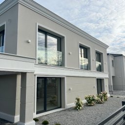 FUTURO HOUSE SPÓŁKA Z OGRANICZONĄ ODPOWIEDZIALNOŚCIĄ - Elegancki, dwukondygnacyjny dom z szarą elewacją, białymi zdobieniami i dużymi oknami. Uwagę przykuwają kwiaty przed budynkiem i kostka brukowa.