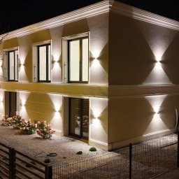 FUTURO HOUSE SPÓŁKA Z OGRANICZONĄ ODPOWIEDZIALNOŚCIĄ - Dwukondygnacyjny dom z elewacją w odcieniach beżu, oświetlony lampami ściennymi, z widocznym fragmentem ogrodu i ogrodzenia, nocna sceneria.