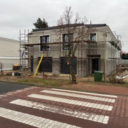 FUTURO HOUSE SPÓŁKA Z OGRANICZONĄ ODPOWIEDZIALNOŚCIĄ - Szary dom w trakcie budowy, otoczony rusztowaniami i drzewem bez liści. Widok z poziomu ulicy, z przejściem dla pieszych na pierwszym planie. Pochmurne niebo.