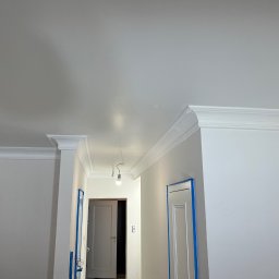 FUTURO HOUSE SPÓŁKA Z OGRANICZONĄ ODPOWIEDZIALNOŚCIĄ - Białe wnętrze po malowaniu z dekoracyjnymi listwami przysufitowymi i zabezpieczonymi taśmą malarską ościeżnicami drzwi, widok na korytarz z oświetleniem.