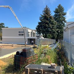 FUTURO HOUSE SPÓŁKA Z OGRANICZONĄ ODPOWIEDZIALNOŚCIĄ - Wylewka betonowa przed nowoczesnym domem z użyciem pompy do betonu. Na pierwszym planie paleta z dachówkami. Słoneczny dzień, błękitne niebo.