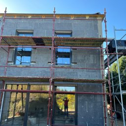 FUTURO HOUSE SPÓŁKA Z OGRANICZONĄ ODPOWIEDZIALNOŚCIĄ - Dom w trakcie ocieplania, widoczne rusztowania i szara izolacja. W oknach odbija się osoba robiąca zdjęcie. Częściowo ocieplona elewacja budynku.