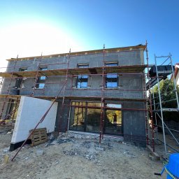 FUTURO HOUSE SPÓŁKA Z OGRANICZONĄ ODPOWIEDZIALNOŚCIĄ - Budowa domu w trakcie: ocieplony budynek z rusztowaniami, widoczne okna i fragment ściany z białych bloczków. W tle błękitne niebo i sąsiedni dom.