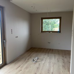 FUTURO HOUSE SPÓŁKA Z OGRANICZONĄ ODPOWIEDZIALNOŚCIĄ - Jasne panele podłogowe w pokoju z beżowymi ścianami, widoczne puszki elektryczne, okno z widokiem na zieleń i kabel od oświetlenia sufitowego. Wykończenie wnętrz w nowym domu.