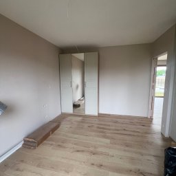 FUTURO HOUSE SPÓŁKA Z OGRANICZONĄ ODPOWIEDZIALNOŚCIĄ - Jasne panele podłogowe w pokoju z beżowymi ścianami i białą szafą z lustrem. Widoczne niedokończone detale, np. wystające kable i listwy przypodłogowe.