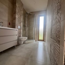 FUTURO HOUSE SPÓŁKA Z OGRANICZONĄ ODPOWIEDZIALNOŚCIĄ - Jasna łazienka z kabiną prysznicową, umywalką z szafką i toaletą podwieszaną. Ściany wyłożone płytkami z wzorem, podłoga z dużych, jasnych płytek. Okno w tle.