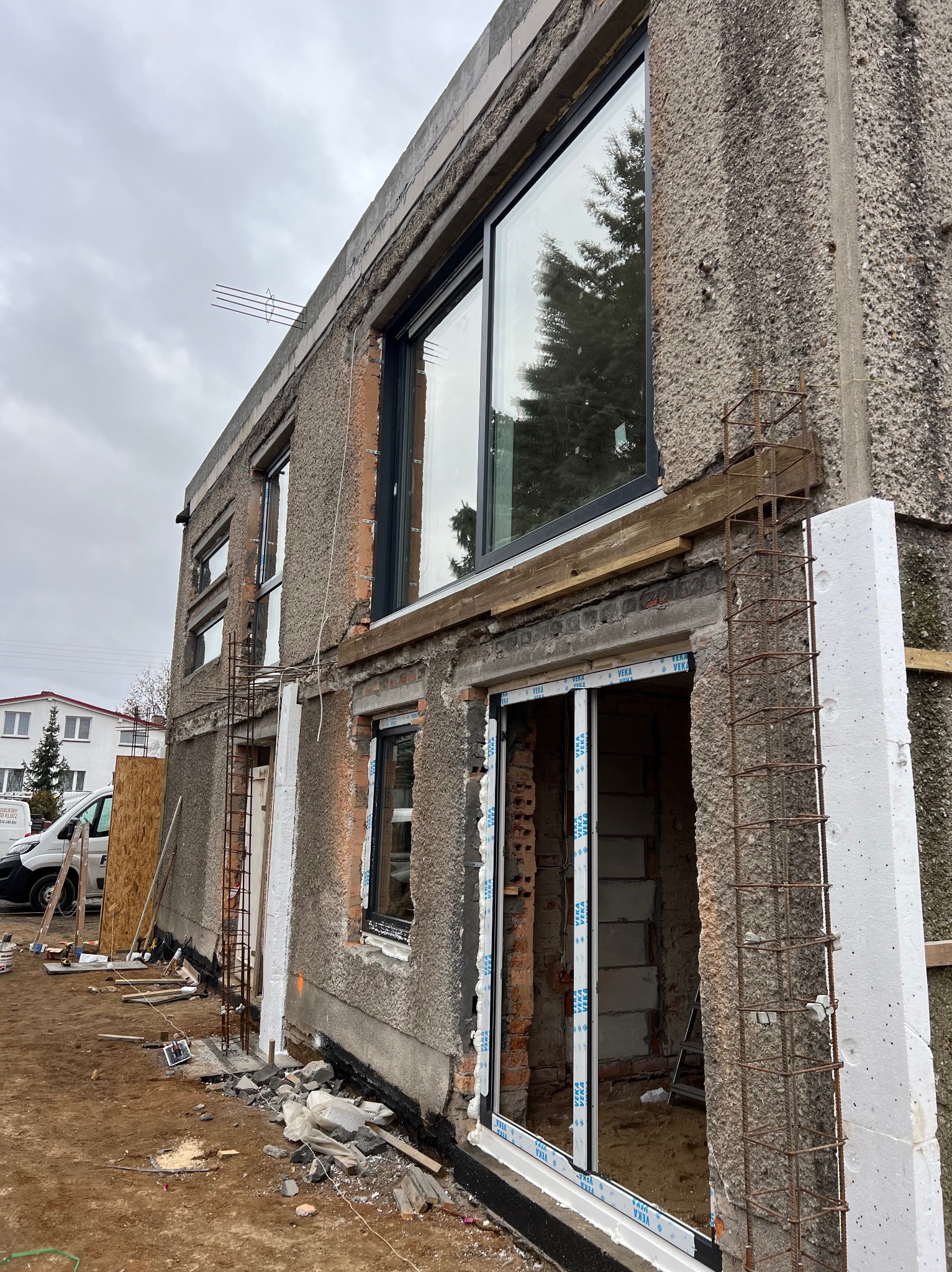 Budynek w trakcie rozbudowy z nowymi oknami i ociepleniem. Widoczne zbrojenia i styropian. Częściowo odsłonięta cegła. Na ziemi gruz i narzędzia.