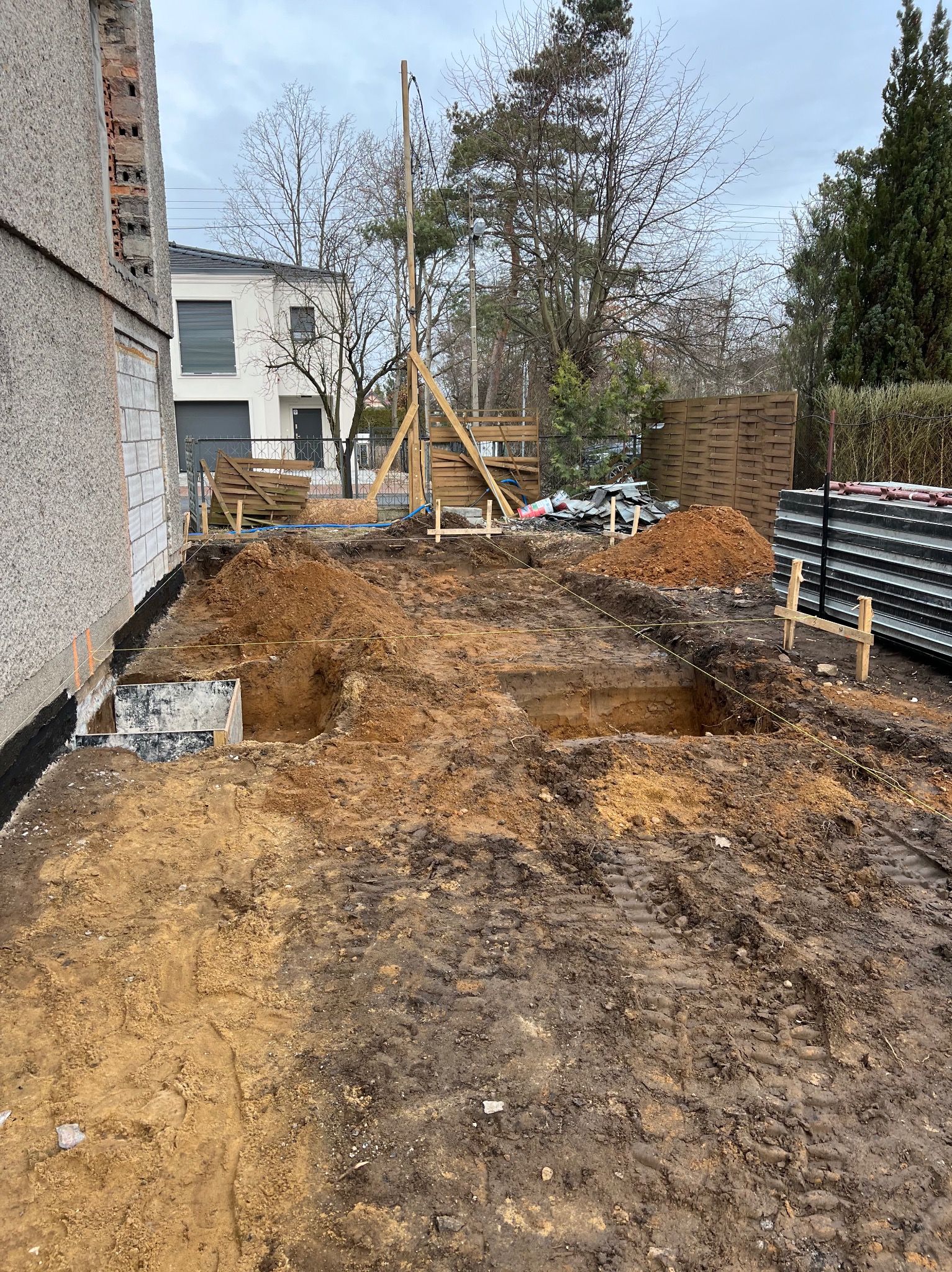 Wykop pod fundamenty domu jednorodzinnego, widoczne szalunki i zabezpieczenie przeciwwilgociowe, w tle budynek mieszkalny i drzewa.