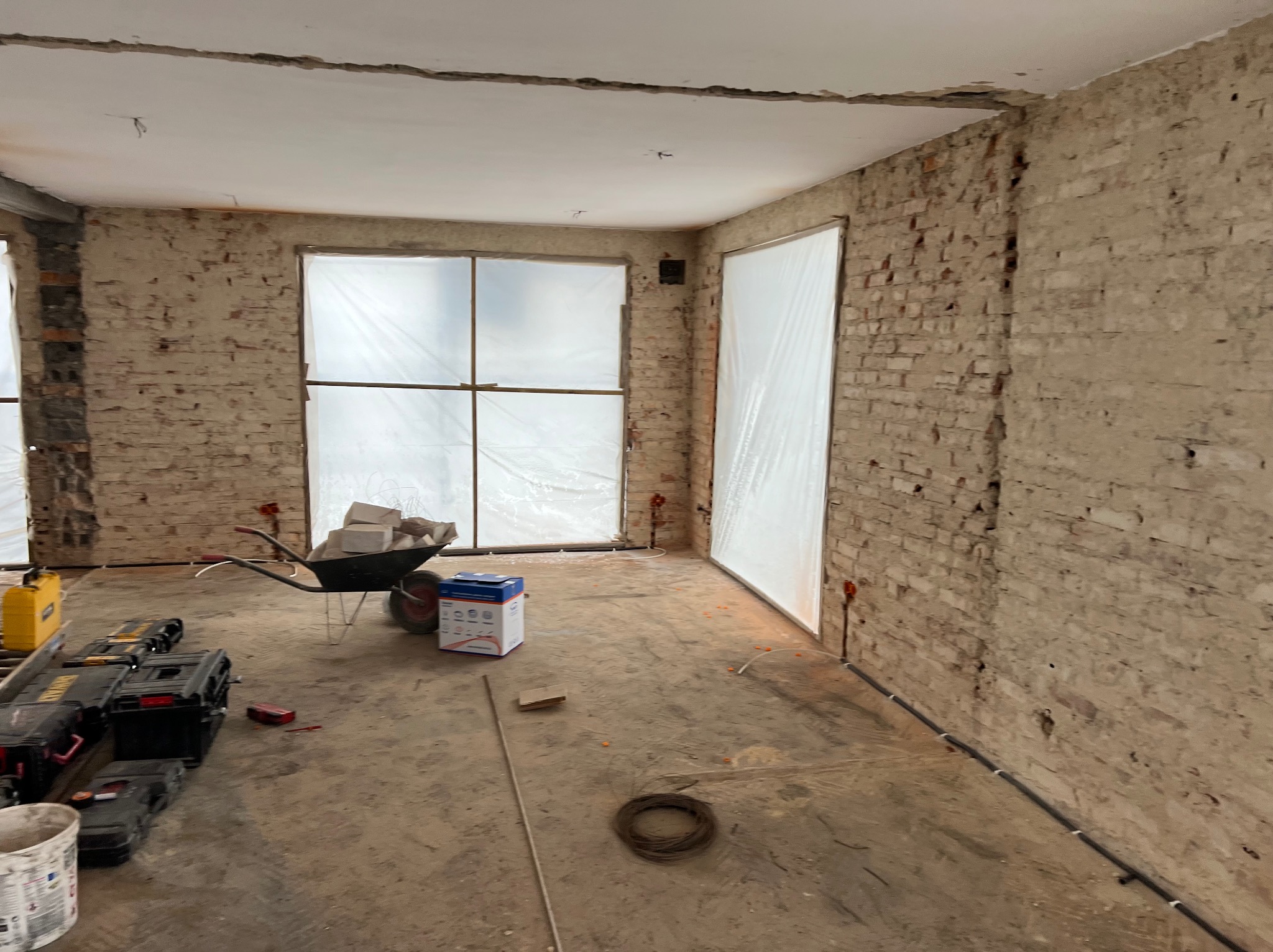 Wnętrze w trakcie remontu: surowe ceglane ściany, okna zabezpieczone folią, taczka z gruzem, narzędzia i materiały budowlane na podłodze. Widoczne ślady po instalacjach.