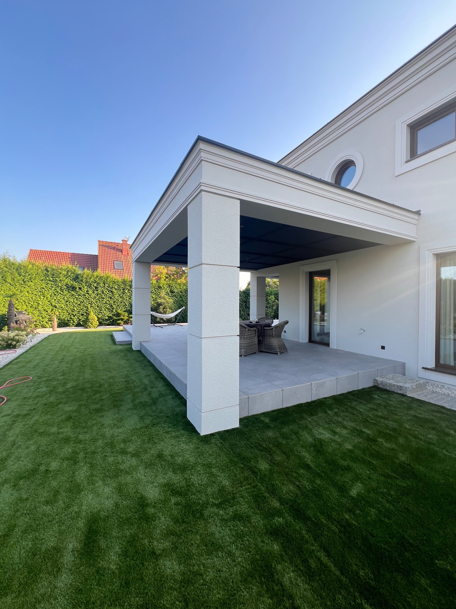 Nowoczesny dom z tarasem i zadbanym trawnikiem. Biała elewacja, geometryczne detale architektoniczne, hamak w tle. Słoneczny dzień, błękitne niebo.