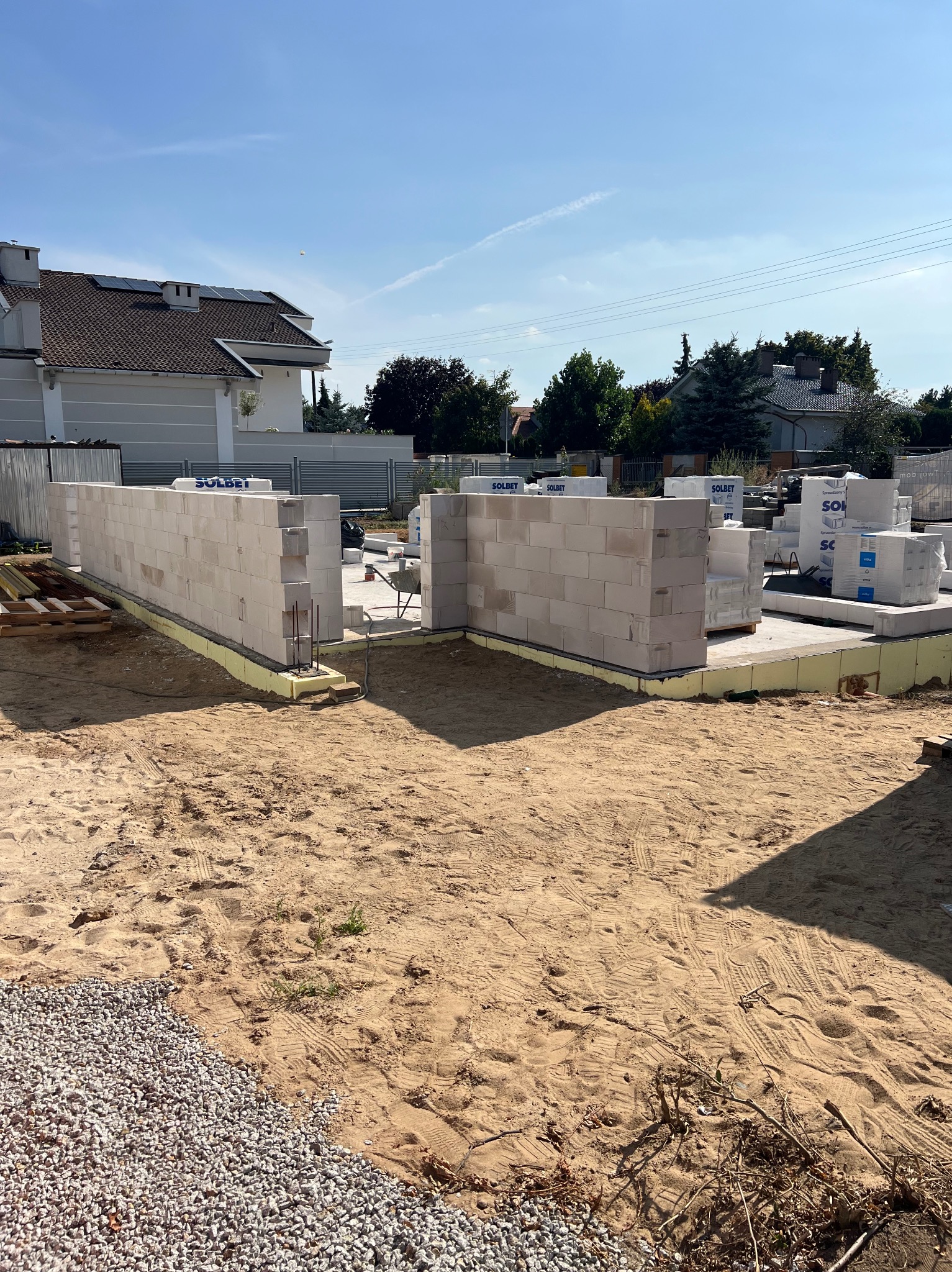 Wznoszenie ścian z białych bloczków Solbet na fundamencie z izolacją. Widoczne materiały budowlane i otoczenie placu budowy w słoneczny dzień.